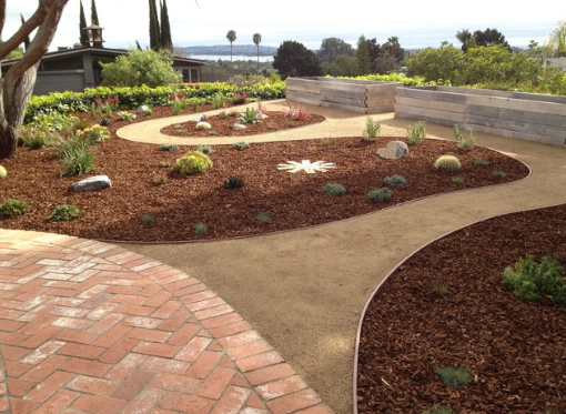 El patio de ladrillo da acceso al jardín con senderos curvos, mantillo y bancales elevados. Vista panorámica al fondo.