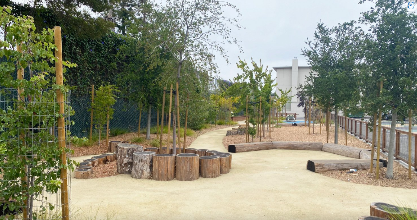 Parque infantil con suelo beige, troncos de madera y árboles. Valla al fondo y cielo nublado.