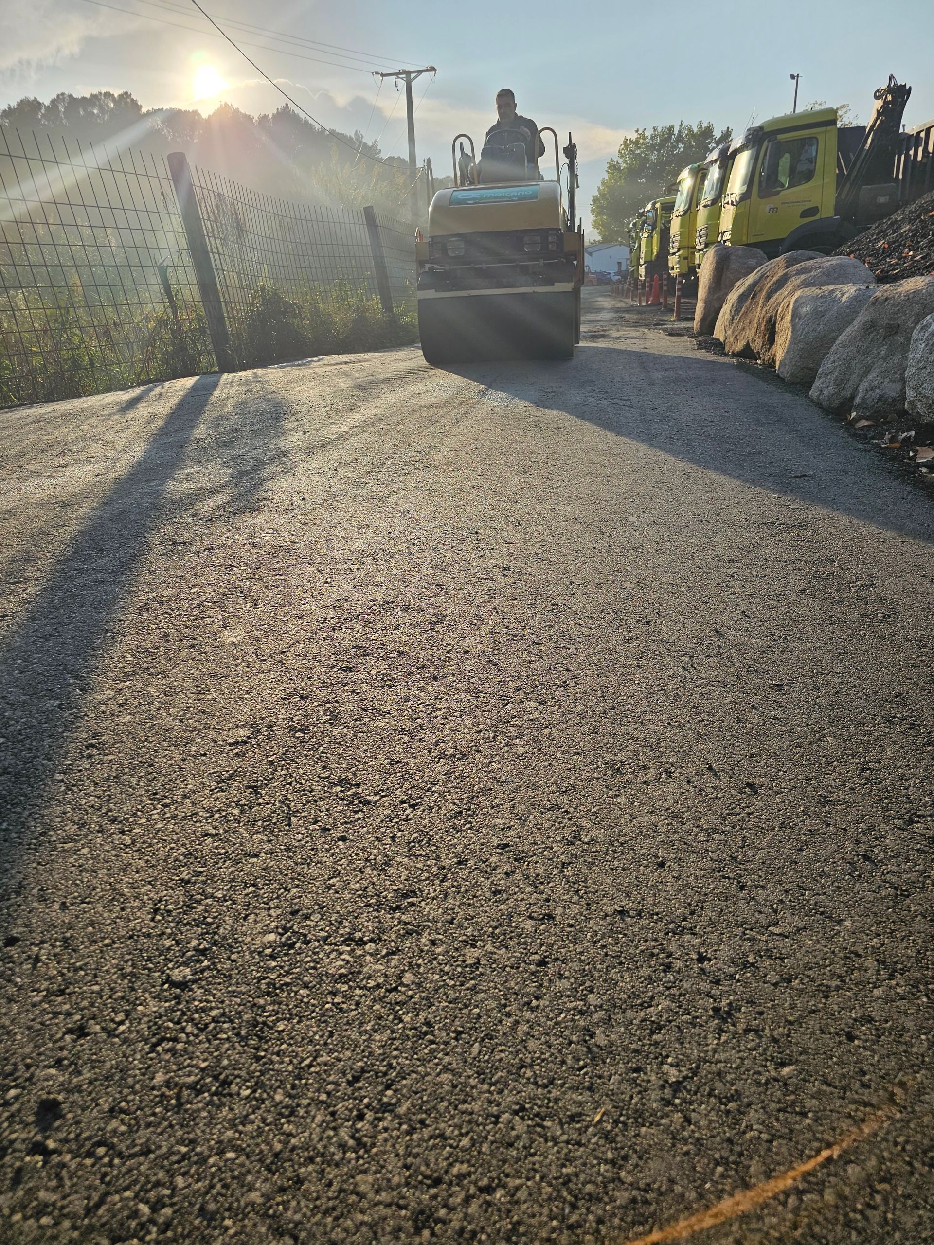 Hombre operando un compactador vibratorio en un camino de grava, con equipo de construcción al fondo.