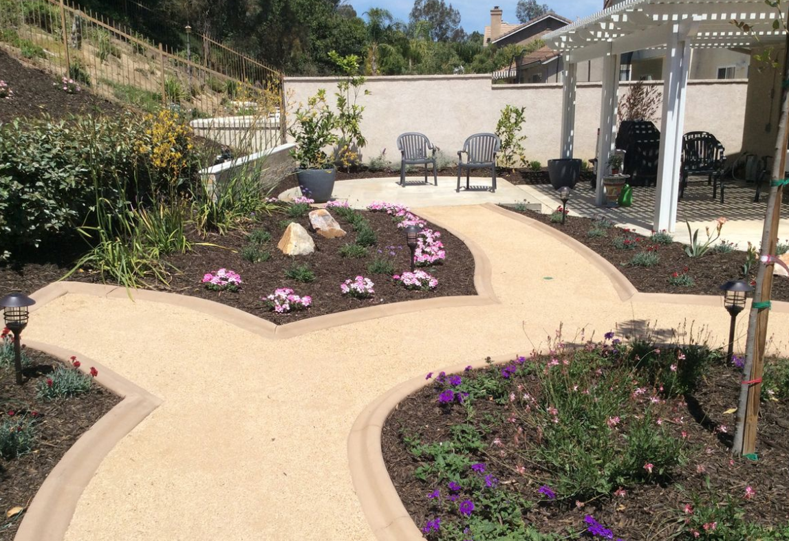 Jardín con senderos de color canela, parterres de flores y una pérgola blanca en un entorno exterior soleado.
