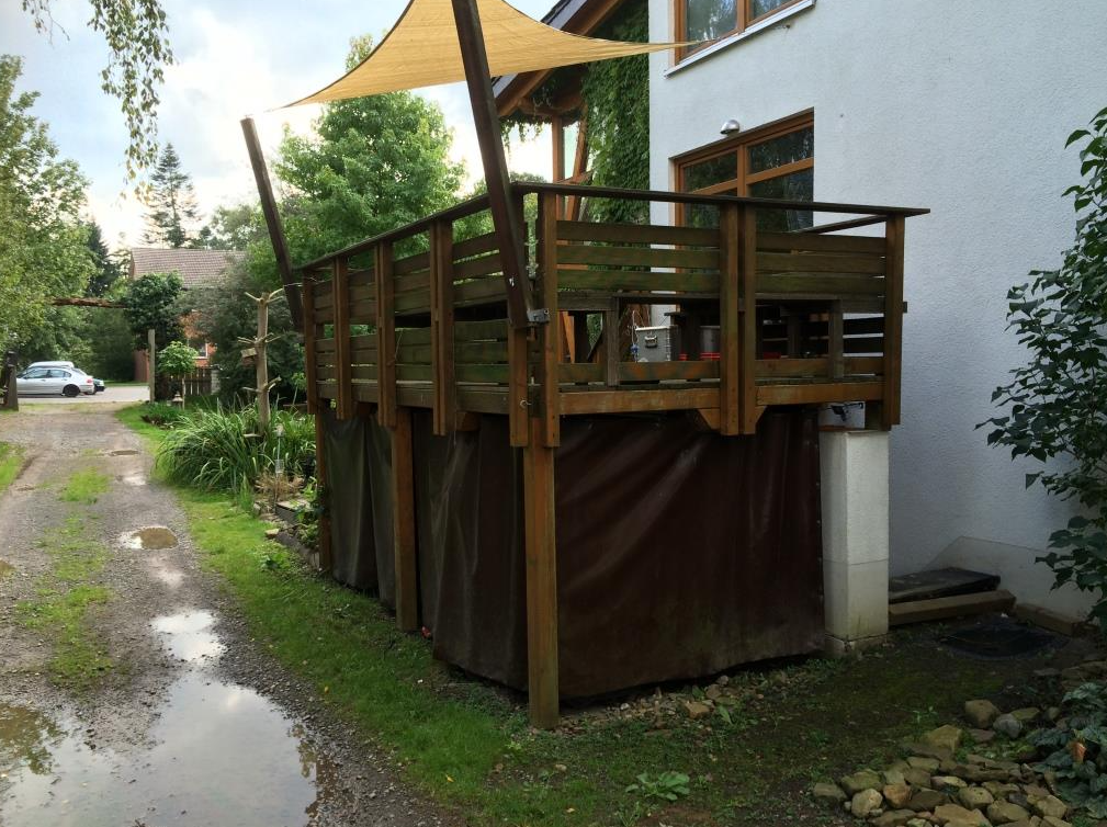 Terraza de madera con vela de sombra, adosada a una casa. Madera marrón, zócalo de lona marrón y césped verde.