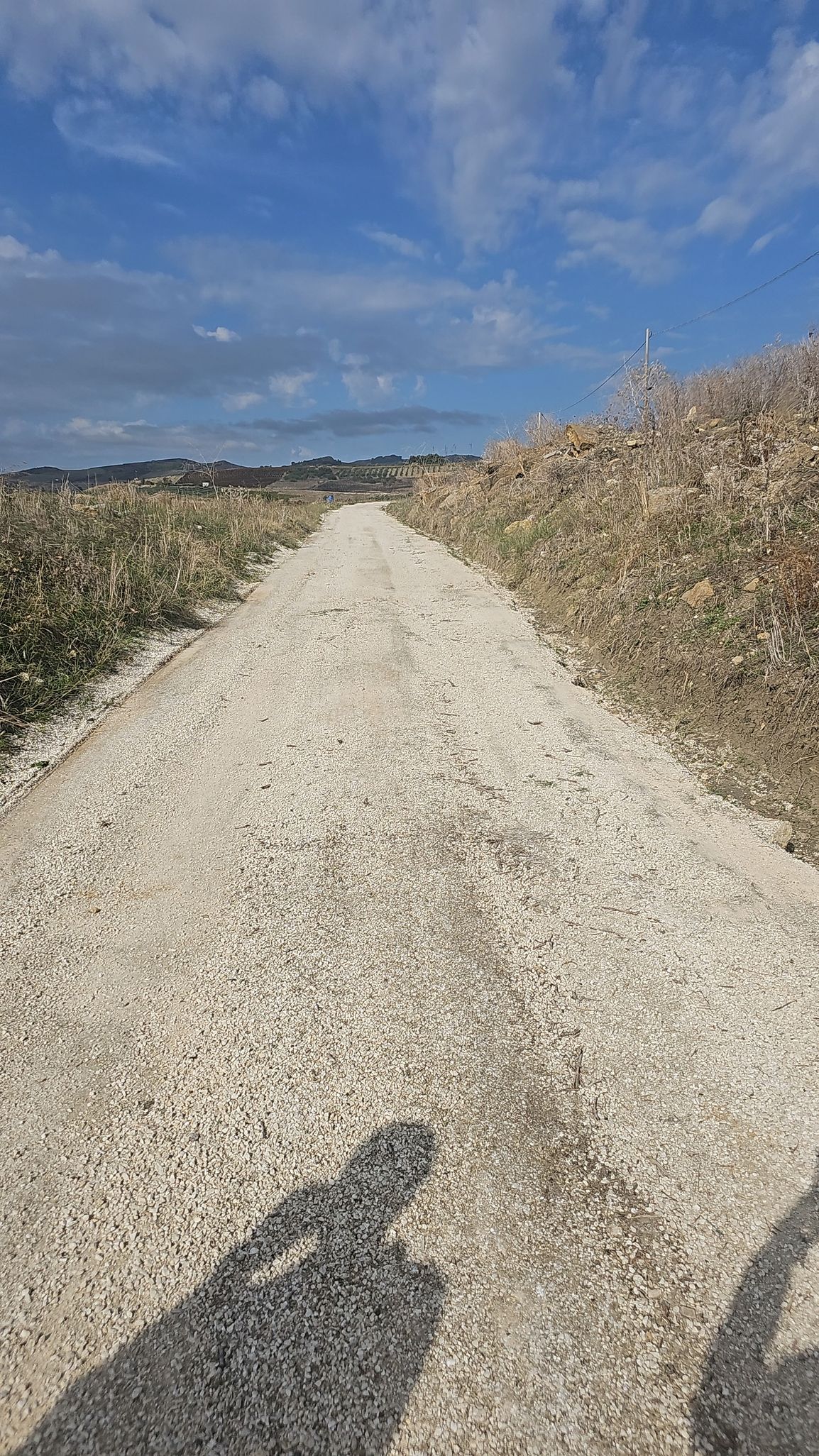 Camino de grava entre campos, bajo un cielo azul con nubes.