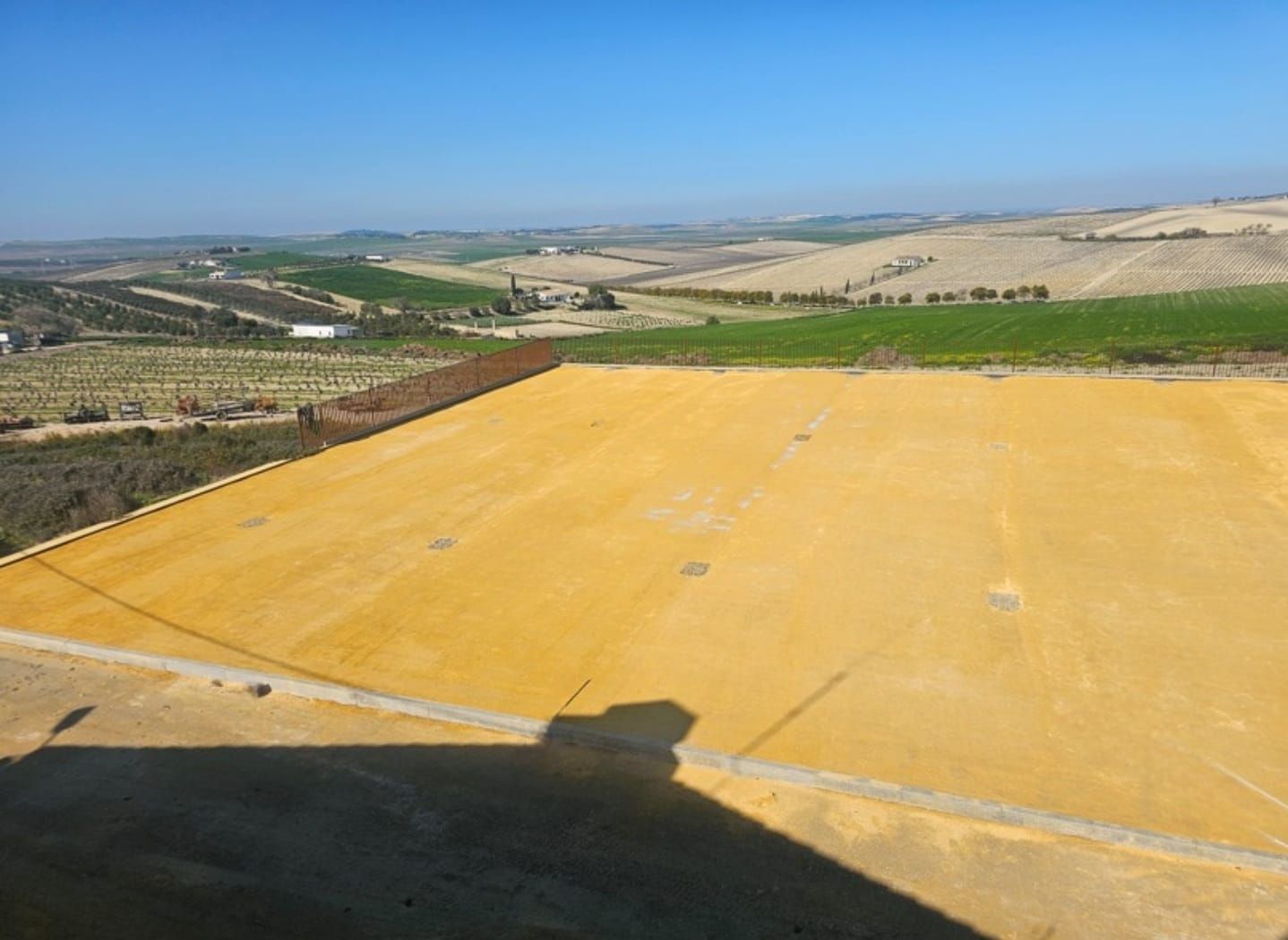 Una extensa estructura cuadrada de color amarillo anaranjado en la cima de una colina con vistas a un paisaje rural y un cielo azul.