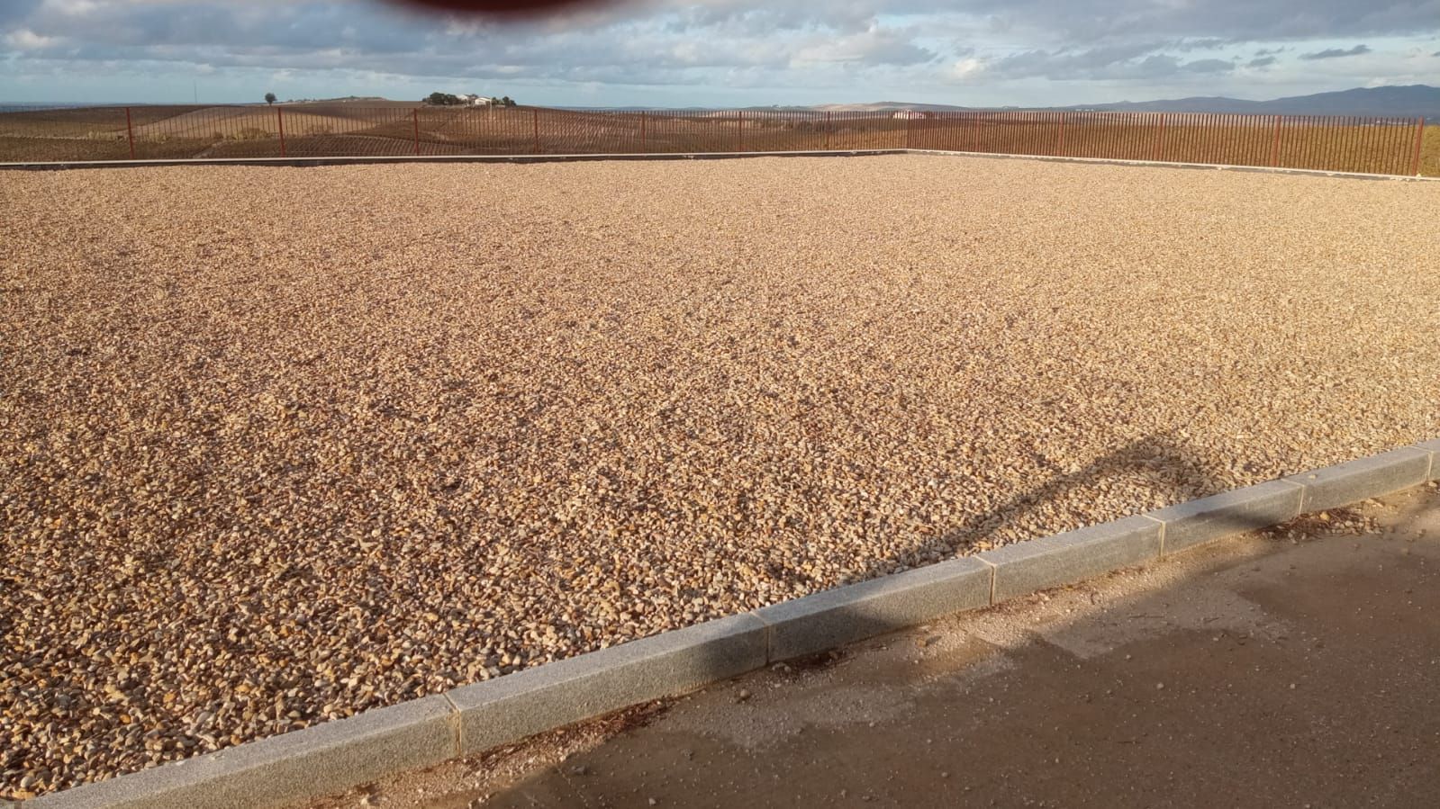 Solar cubierto de grava con bordillo y valla de hormigón al fondo, bajo un cielo parcialmente nublado.