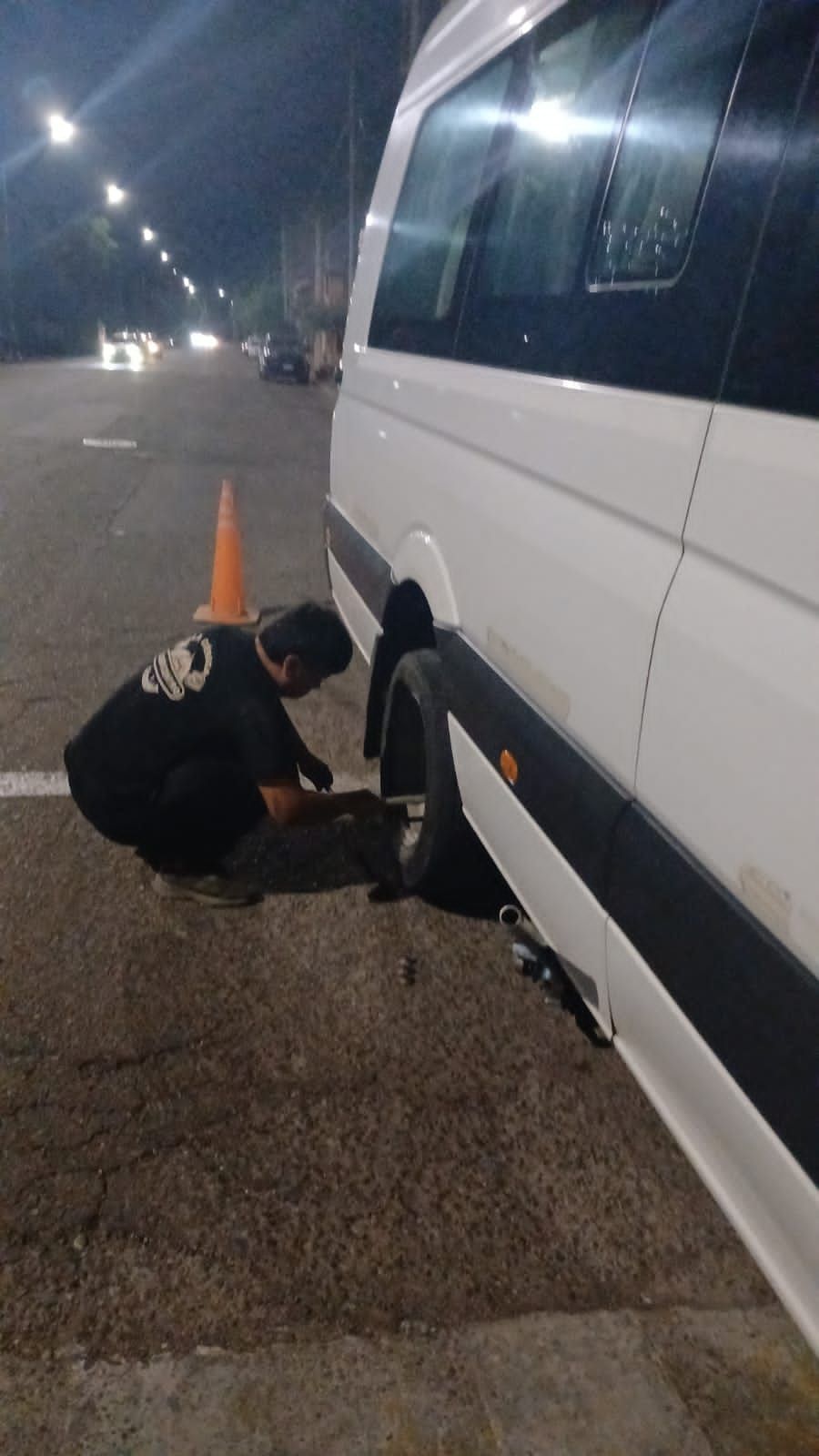 Una persona se agacha, cambiando una llanta de una camioneta blanca por la noche. Cerca hay un cono de tráfico.