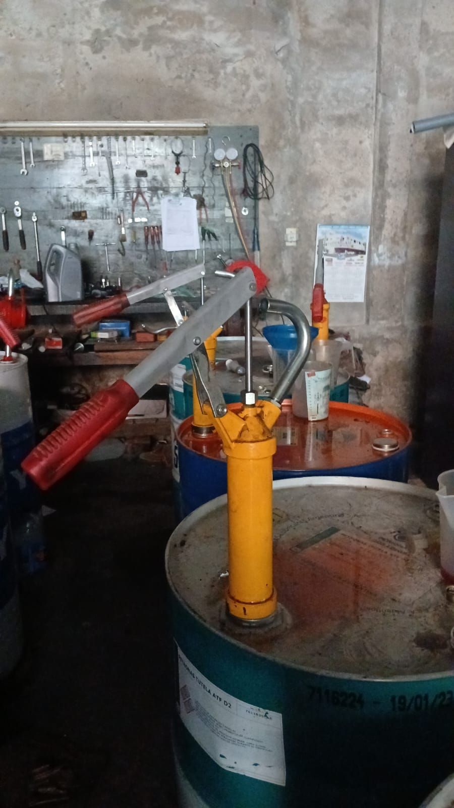 Bomba amarilla sobre un tambor en un taller; mango rojo, fondo de metal, herramientas en la pared.