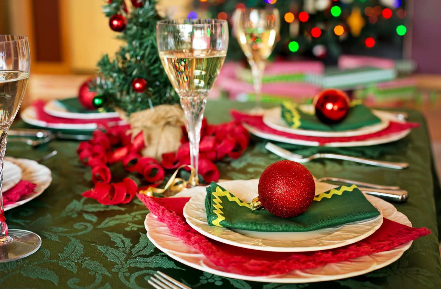 A christmas table setting with plates , wine glasses , and a christmas tree in the background.