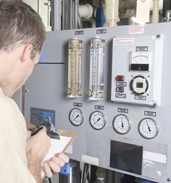 A person in a light-colored shirt records readings from gauges and flow meters on a gray industrial control panel.
