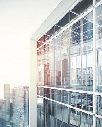 A low-angle view of a modern glass skyscraper with reflections of nearby city buildings against a bright, hazy sky.