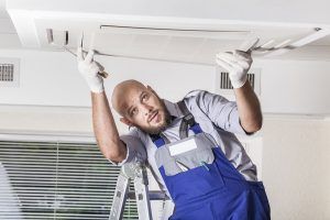 A technician in blue overalls and white gloves stands on a ladder, installing a ceiling-mounted air conditioning unit.