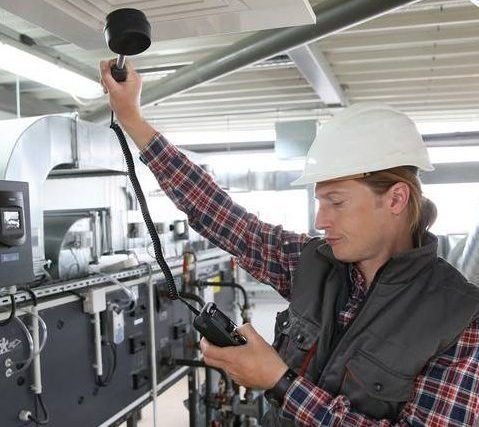 Technician in a hard hat and flannel shirt checks the air output of an HVAC system using a handheld measuring device.