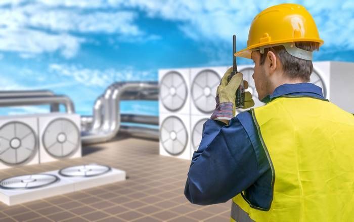 A technician in a yellow hard hat and high-visibility vest holds a two-way radio on a rooftop with HVAC equipment.
