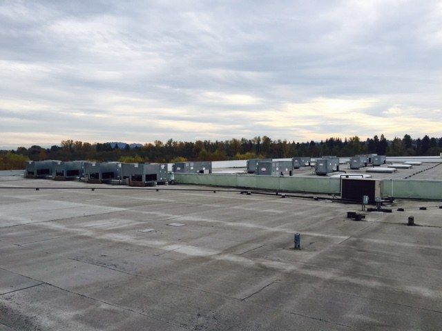 A flat, industrial rooftop under a cloudy sky, featuring multiple large, gray HVAC units lined up in the distance.