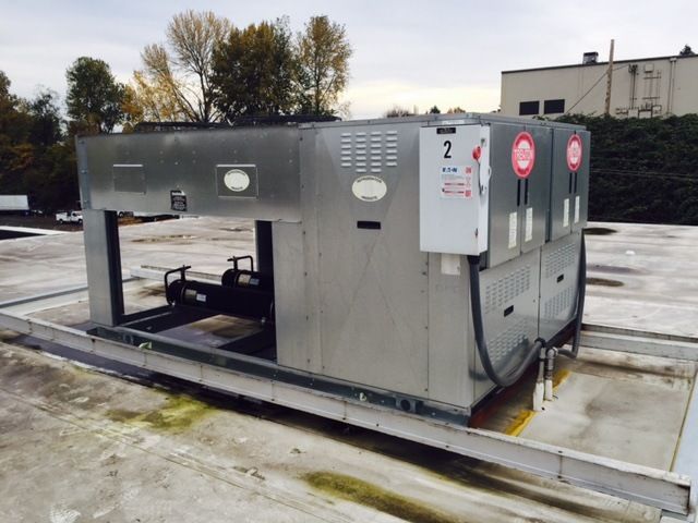 A large industrial HVAC unit sits on a flat building rooftop with a gray metal casing and an attached electrical box.
