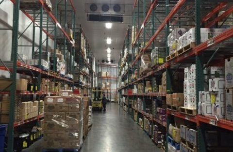 An aisle in a refrigerated warehouse featuring tall metal shelving stocked with boxes and a forklift moving in the distance.