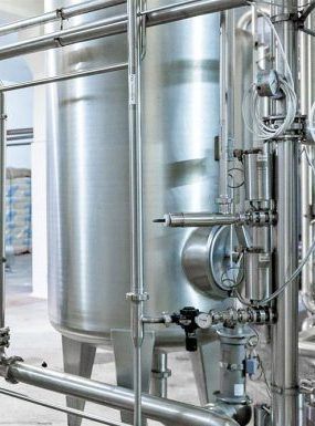 Industrial stainless steel tank with plumbing, valves, and gauges in a manufacturing or processing facility.