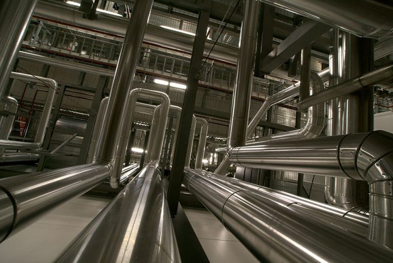 Shiny, metallic industrial pipes arranged in parallel and angular patterns within a dim, mechanical facility.