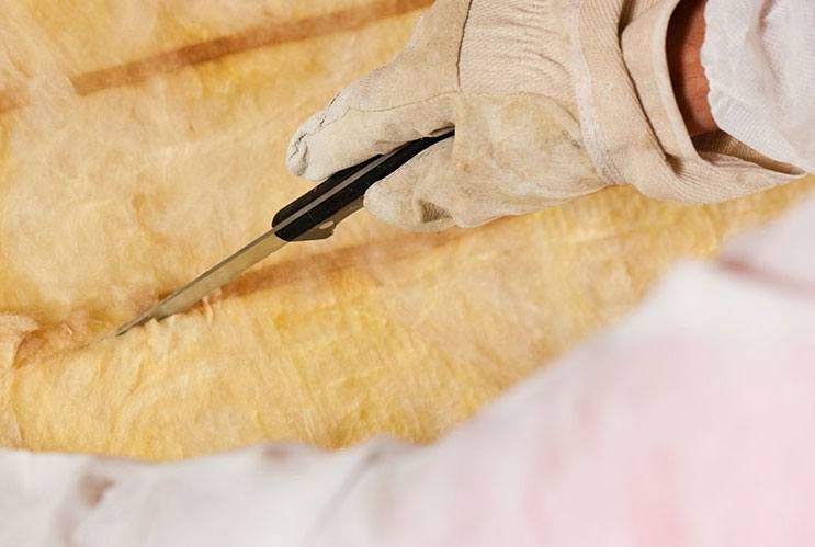 A gloved hand uses a utility knife to cut a yellow fiberglass insulation batt.