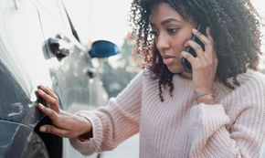 A woman is talking on a cell phone while looking at her car.