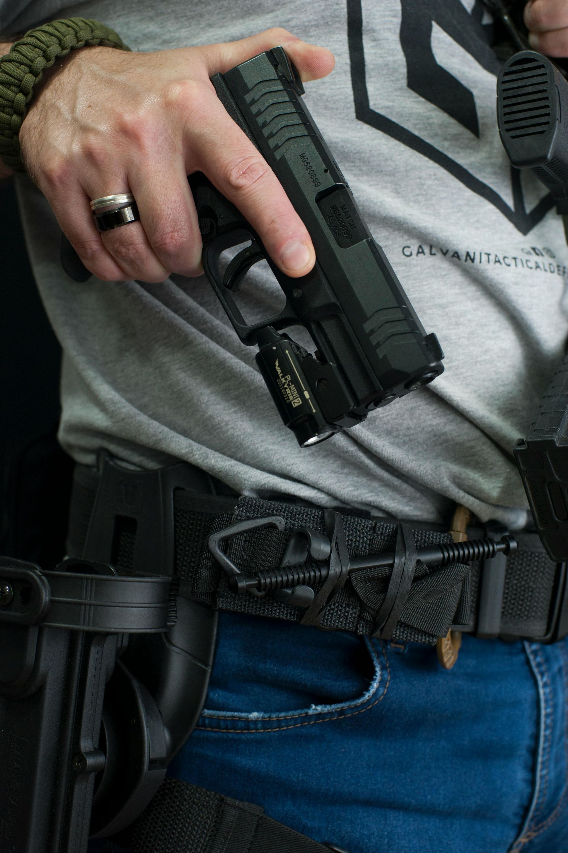 Person holding a handgun with a mounted light, worn on a belt with gear and a tourniquet.