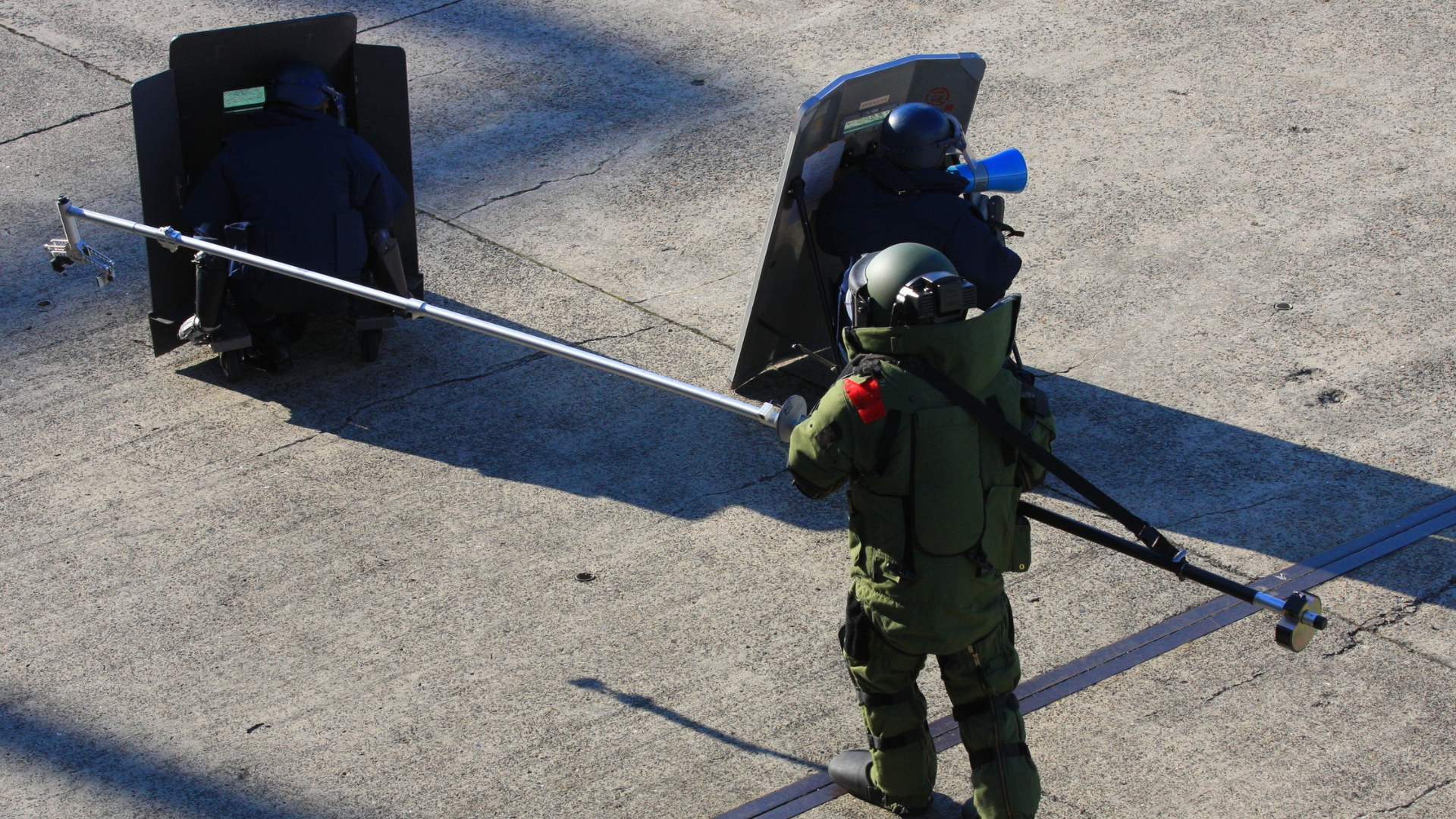Bomb squad officers using an extendable pole to remotely inspect a suspect package behind a protective shield.