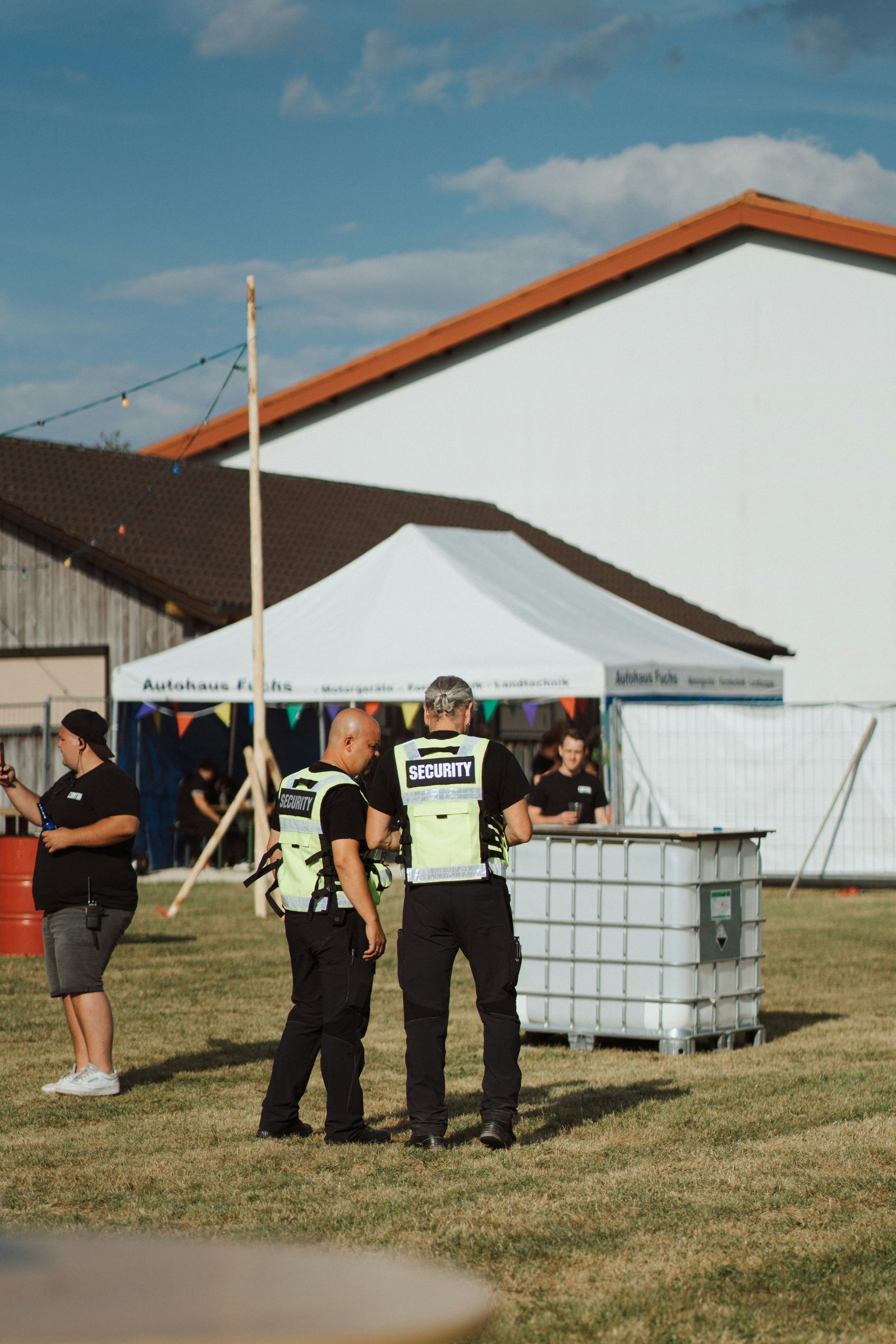Two police officers in vests stand on grass near a white tent and buildings.