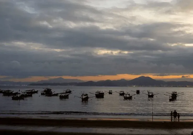 Barcos balançam em águas calmas ao entardecer. Nuvens escuras pairam sobre as montanhas ao longe, com algumas pessoas na praia.