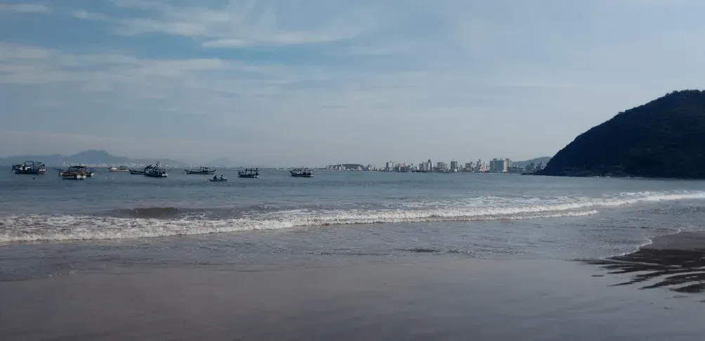 Vista de uma praia com barcos no mar, edifícios no horizonte e uma colina no lado direito sob um céu nublado.