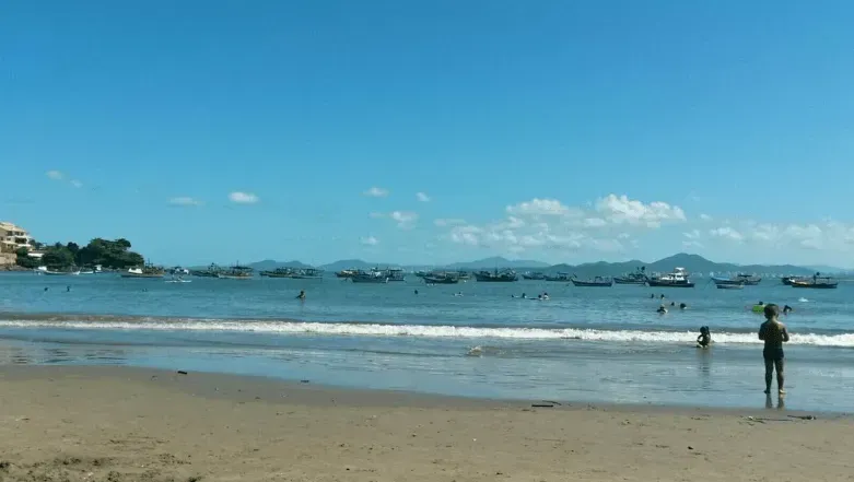 Cena de praia com céu azul, oceano e areia. Vários barcos na água e algumas pessoas nadando. Uma pessoa está em pé perto da beira da água.