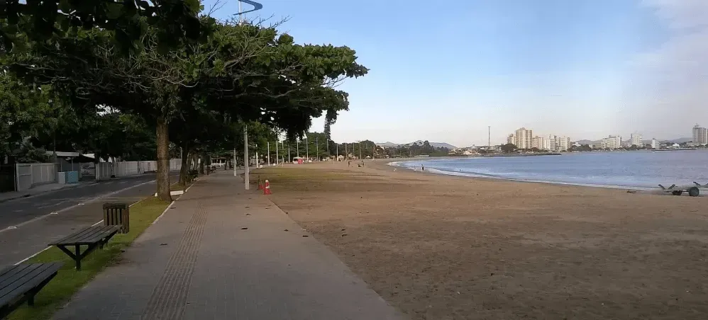 Uma cena de praia com areia, água e árvores. Uma passarela pavimentada com bancos acompanha a praia. Prédios da cidade são visíveis ao longe.