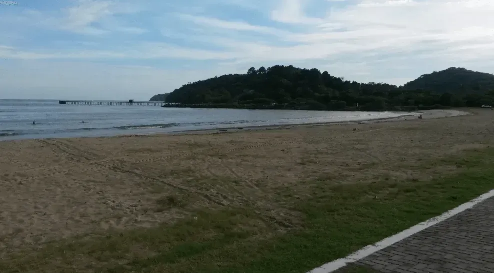 Praia com uma faixa gramada, litoral arenoso, mar calmo e colinas arborizadas ao fundo. Um píer se estende até a água.