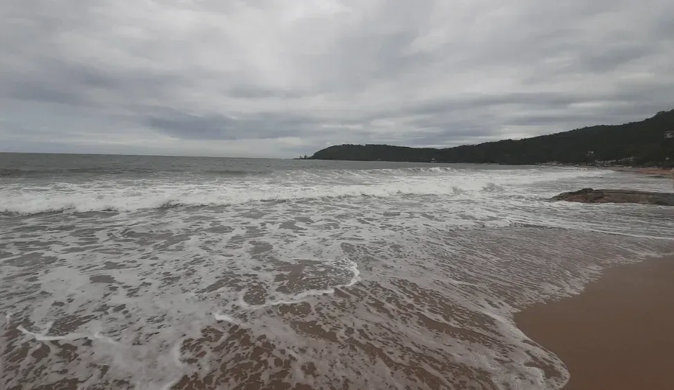 Ondas quebrando em uma praia de areia sob um céu nublado. Ao longe, uma encosta arborizada.