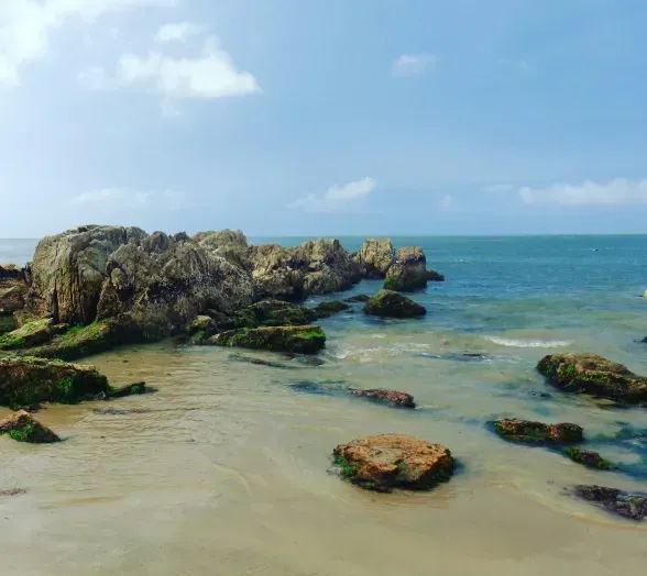 Rochas ao longo de uma praia arenosa, com o oceano ao fundo sob um céu azul com nuvens.