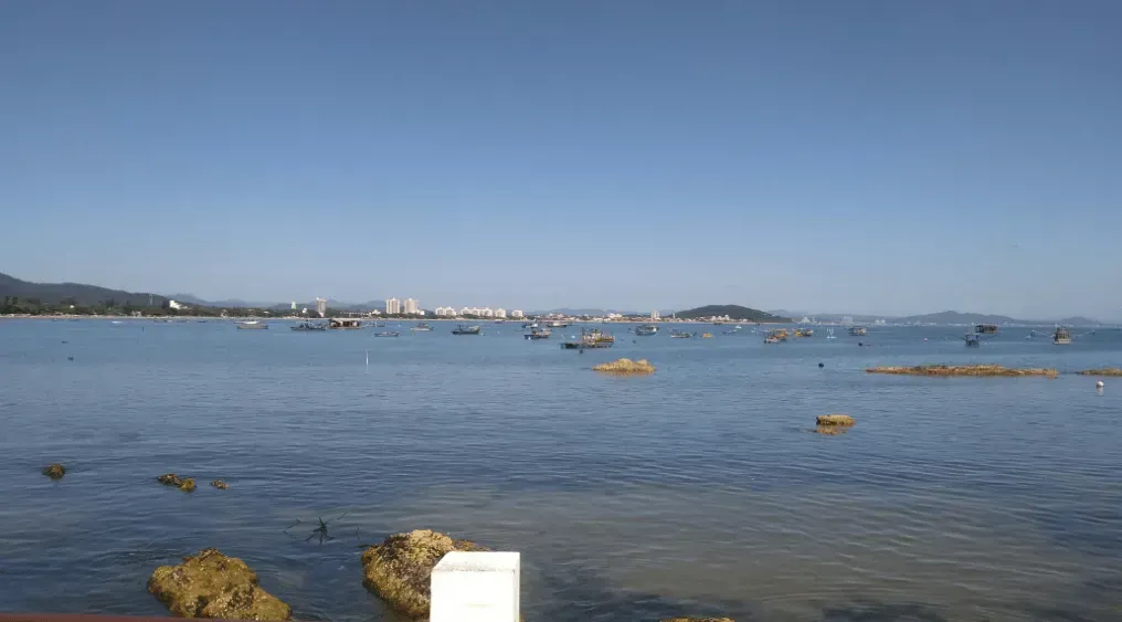 Oceano azul calmo com barcos espalhados e pequenas pedras, com um horizonte de cidade distante sob um céu claro.