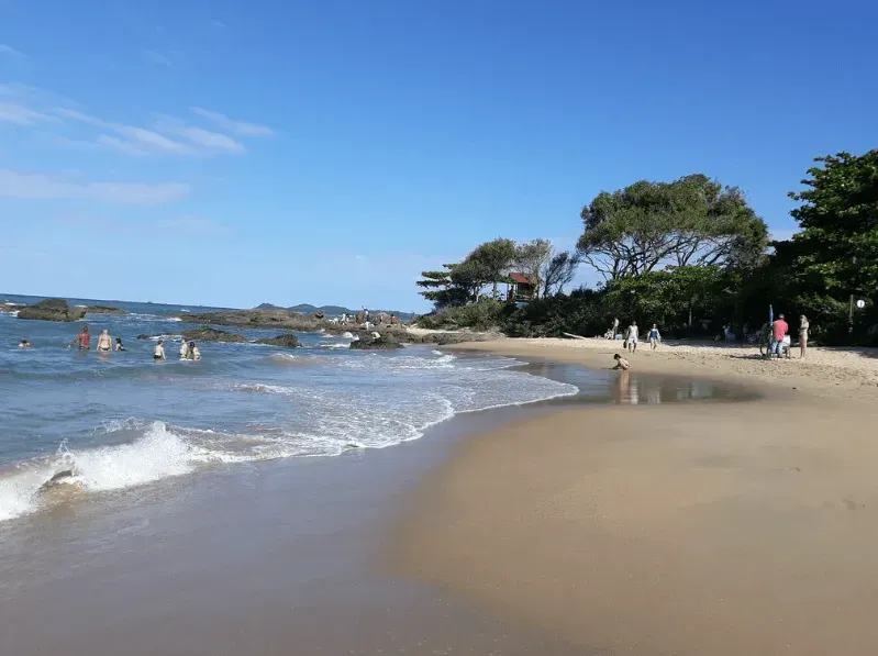 Cena de praia com pessoas nadando e tomando sol sob um céu azul brilhante; ondas quebrando na areia da praia.