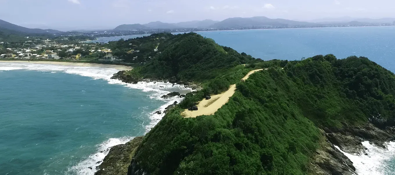 Vista aérea de uma península verdejante projetando-se em águas azul-turquesa, com uma estrada serpenteando ao longo de sua crista. Uma vila margeia a costa próxima.