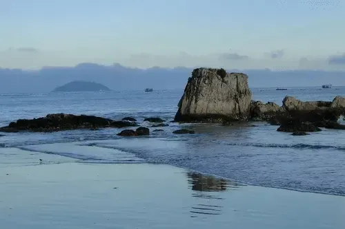 Cena do oceano com grandes formações rochosas, ondas suaves e um horizonte nebuloso sob um céu azul claro.