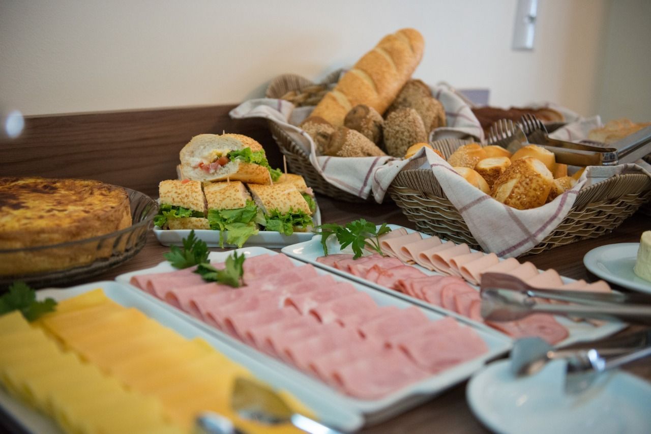 Buffet de café da manhã: presunto, queijo, sanduíches, quiche, pães em cestas.