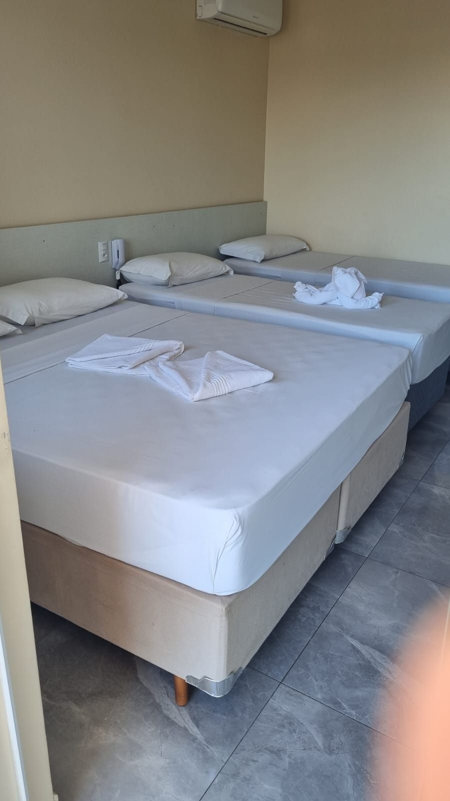 Três camas em um quarto de hotel de cor clara. Lençóis brancos, toalhas dobradas sobre as camas e piso cinza claro.