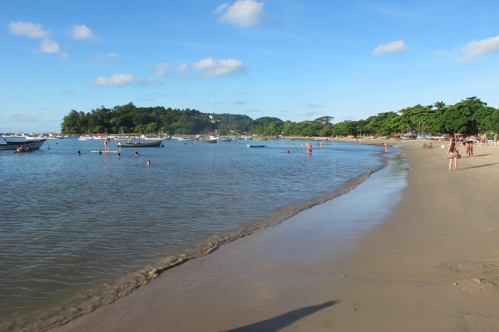 Praia de areia com águas calmas, barcos e pessoas nadando. Árvores e uma pequena ilha ao fundo, sob um céu azul.