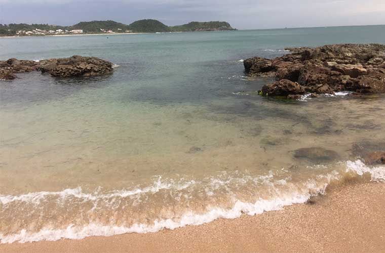 Praia de areia com ondas suaves, afloramentos rochosos e uma costa distante sob um céu nublado.