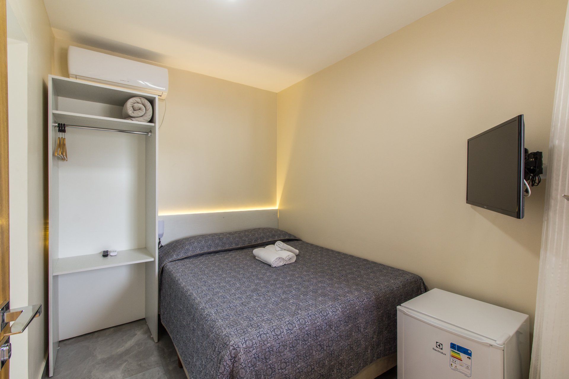 Pequeno quarto de hotel com cama, armário, TV e frigobar. Paredes bege, móveis brancos e colcha cinza.