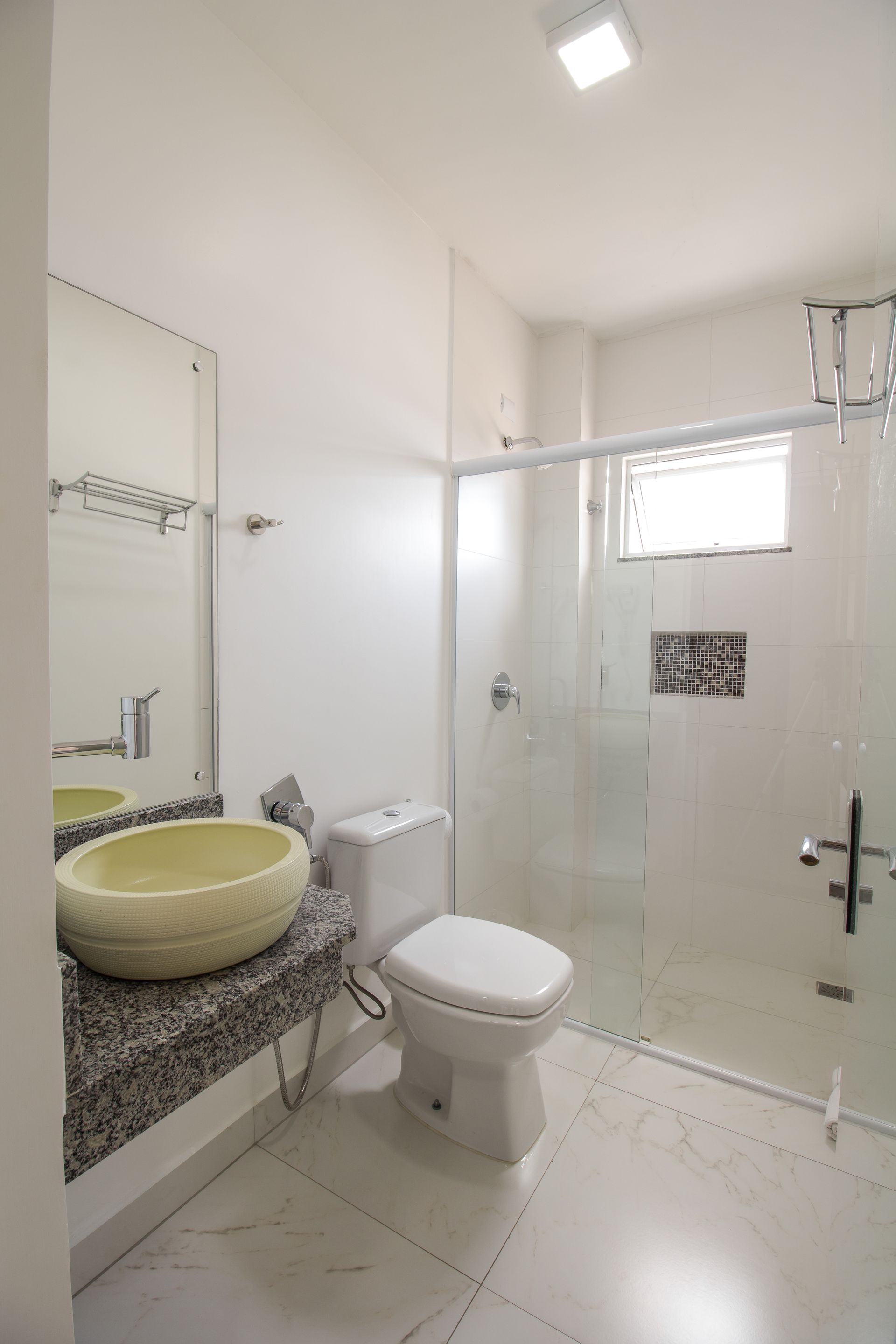 Banheiro branco com bancada de pedra, pia amarela, chuveiro de vidro e vaso sanitário.