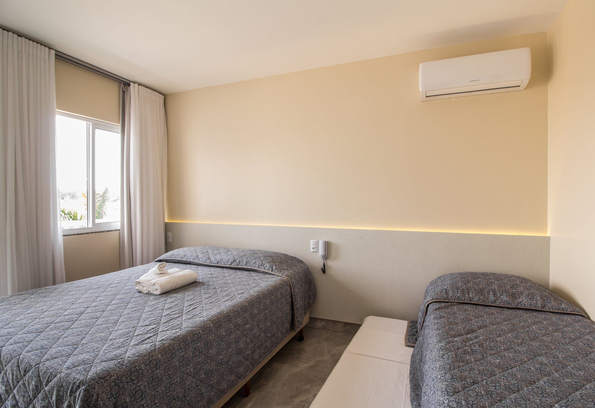 Um quarto de hotel com duas camas de solteiro cobertas com roupa de cama cinza estampada. O quarto tem tons neutros, janela e ar-condicionado.