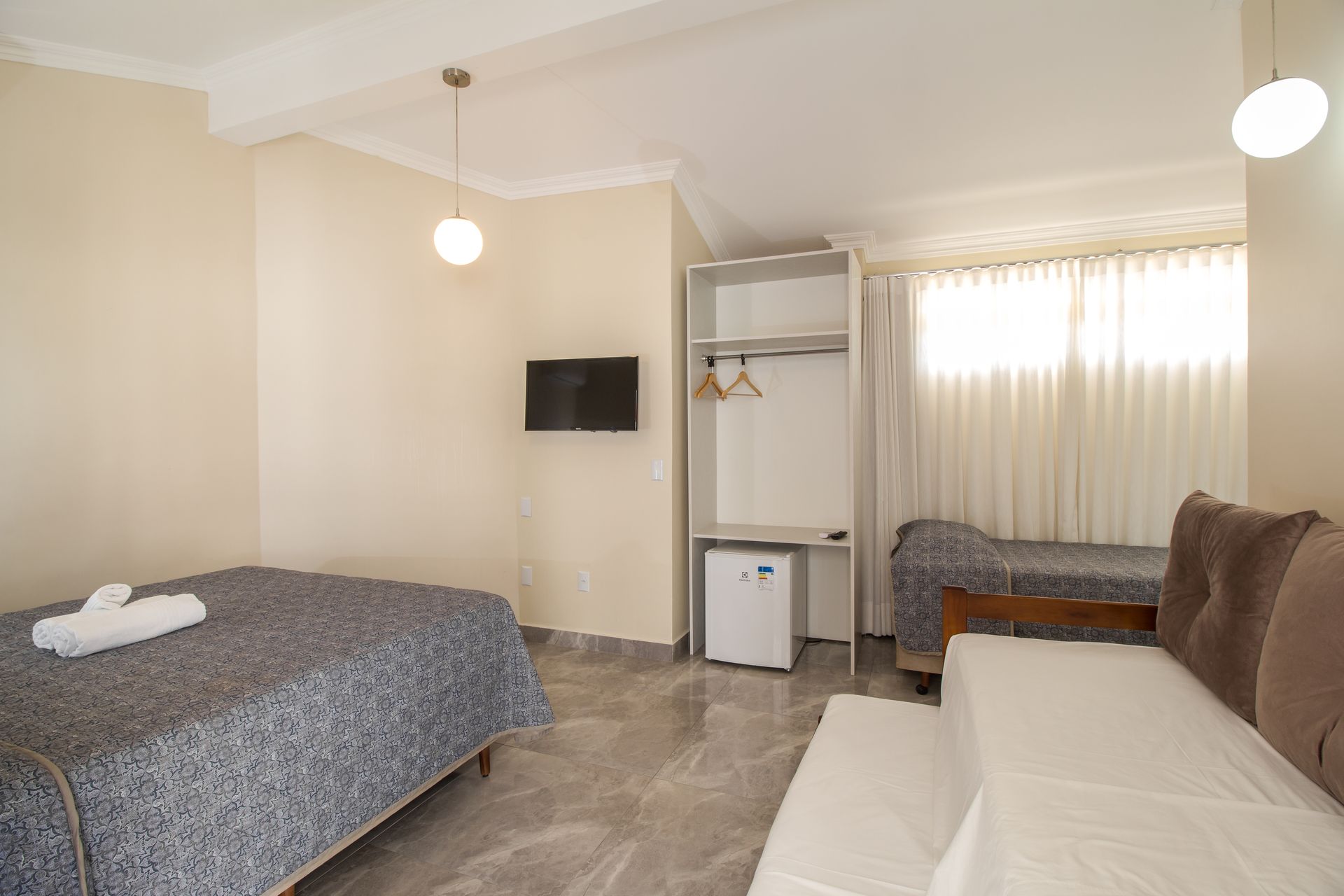 Quarto de hotel com cama, sofá, guarda-roupa, TV e uma pequena geladeira.