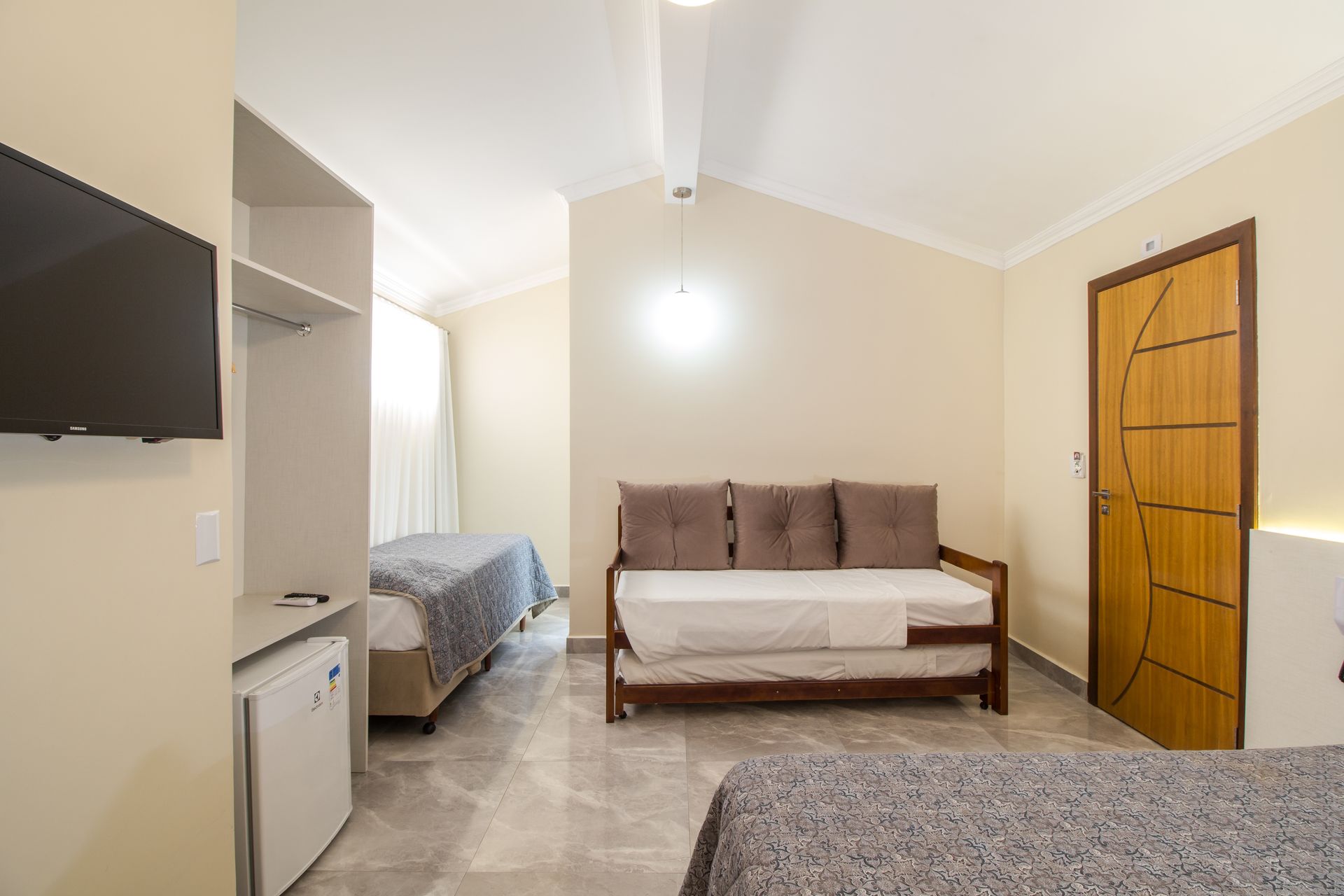 Quarto de hotel com várias camas, TV, sofá e porta. Paredes bege, móveis cinza e marrom.