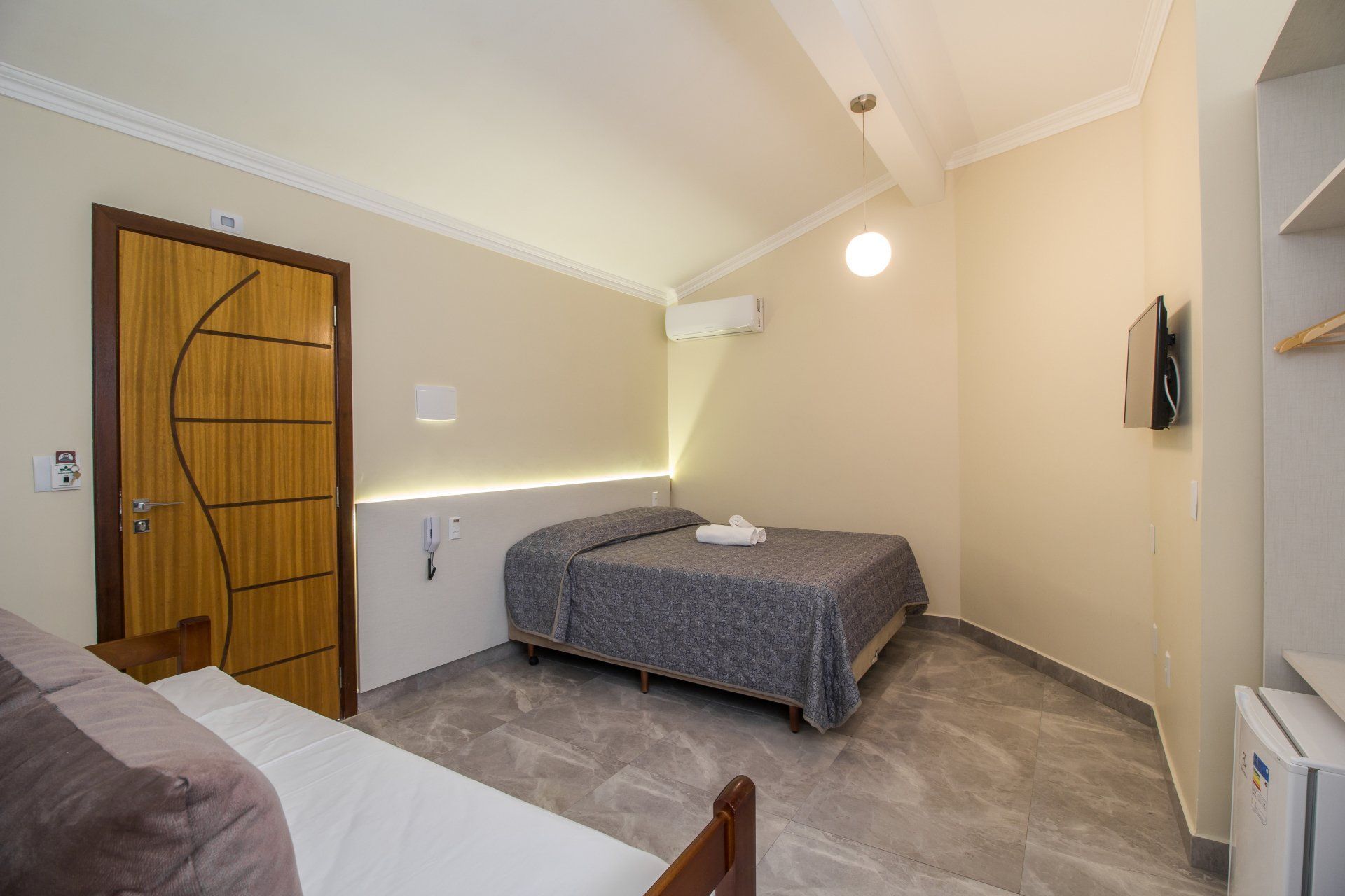Quarto de hotel com cama, sofá-cama e televisão. O quarto tem paredes neutras, piso cinza e porta de madeira.