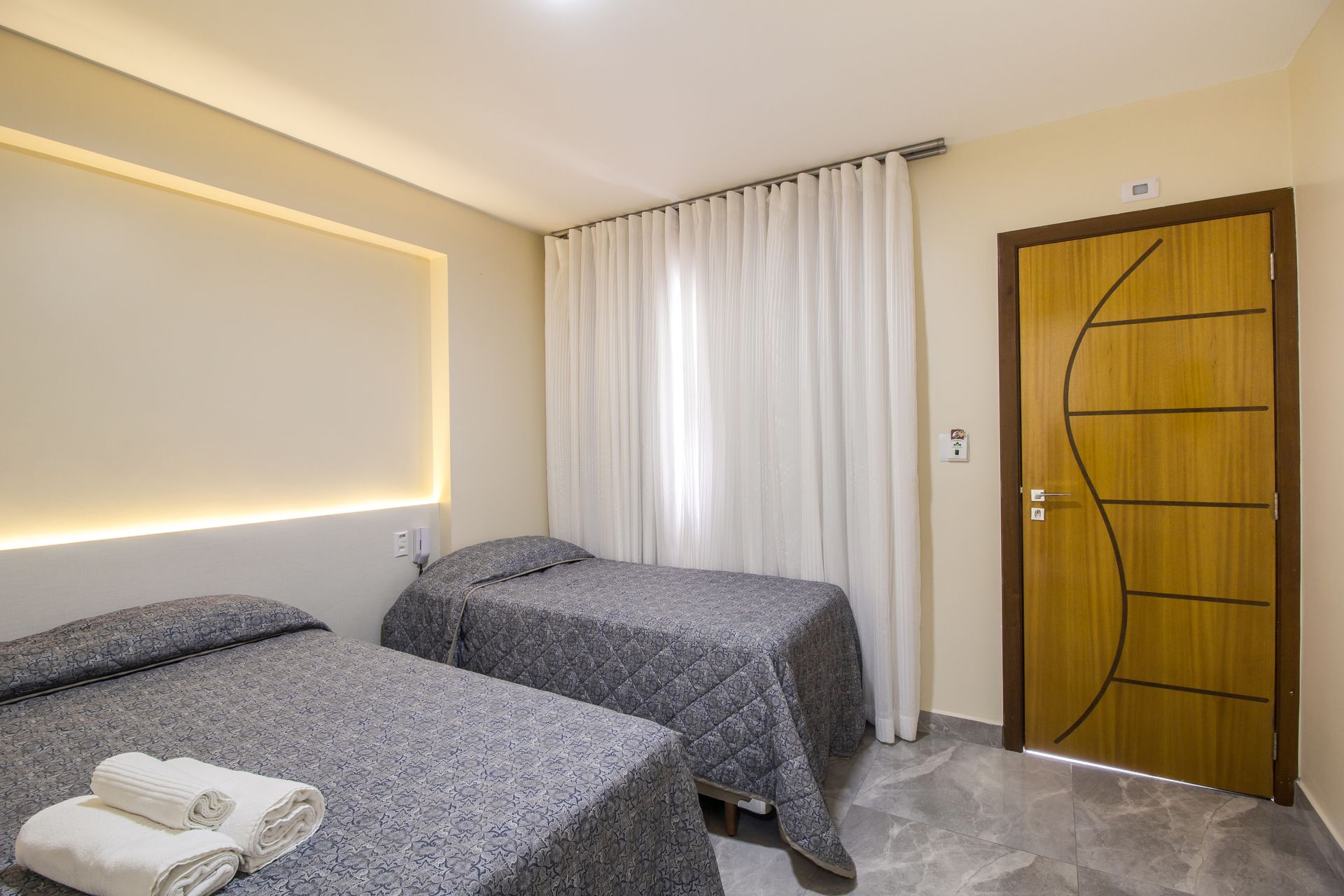 Um pequeno quarto de hotel em tons neutros com duas camas de solteiro, porta fechada e uma janela com cortinas.