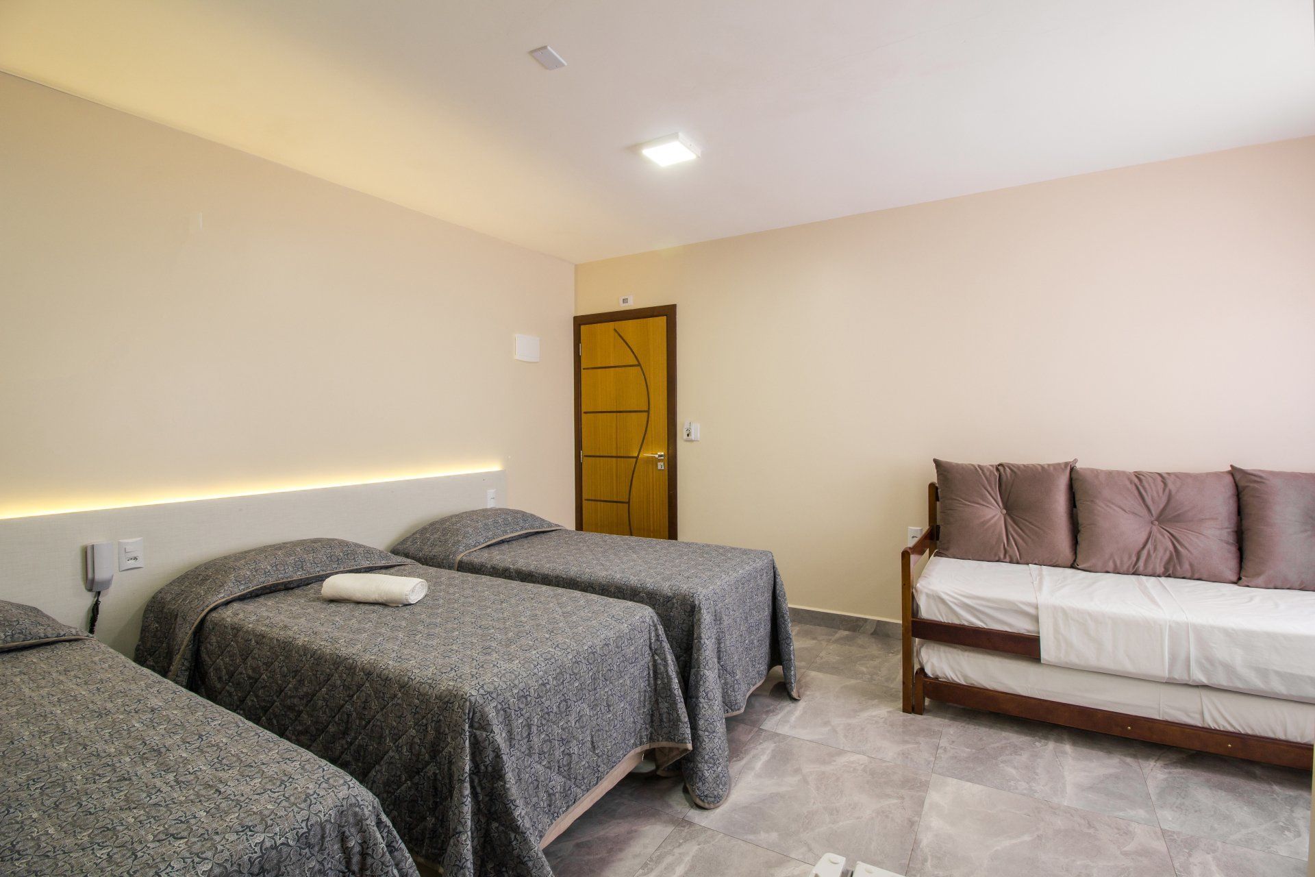 Um quarto de hotel com três camas de solteiro e um sofá. O quarto tem paredes em tons neutros, colchas cinza e uma porta de madeira.