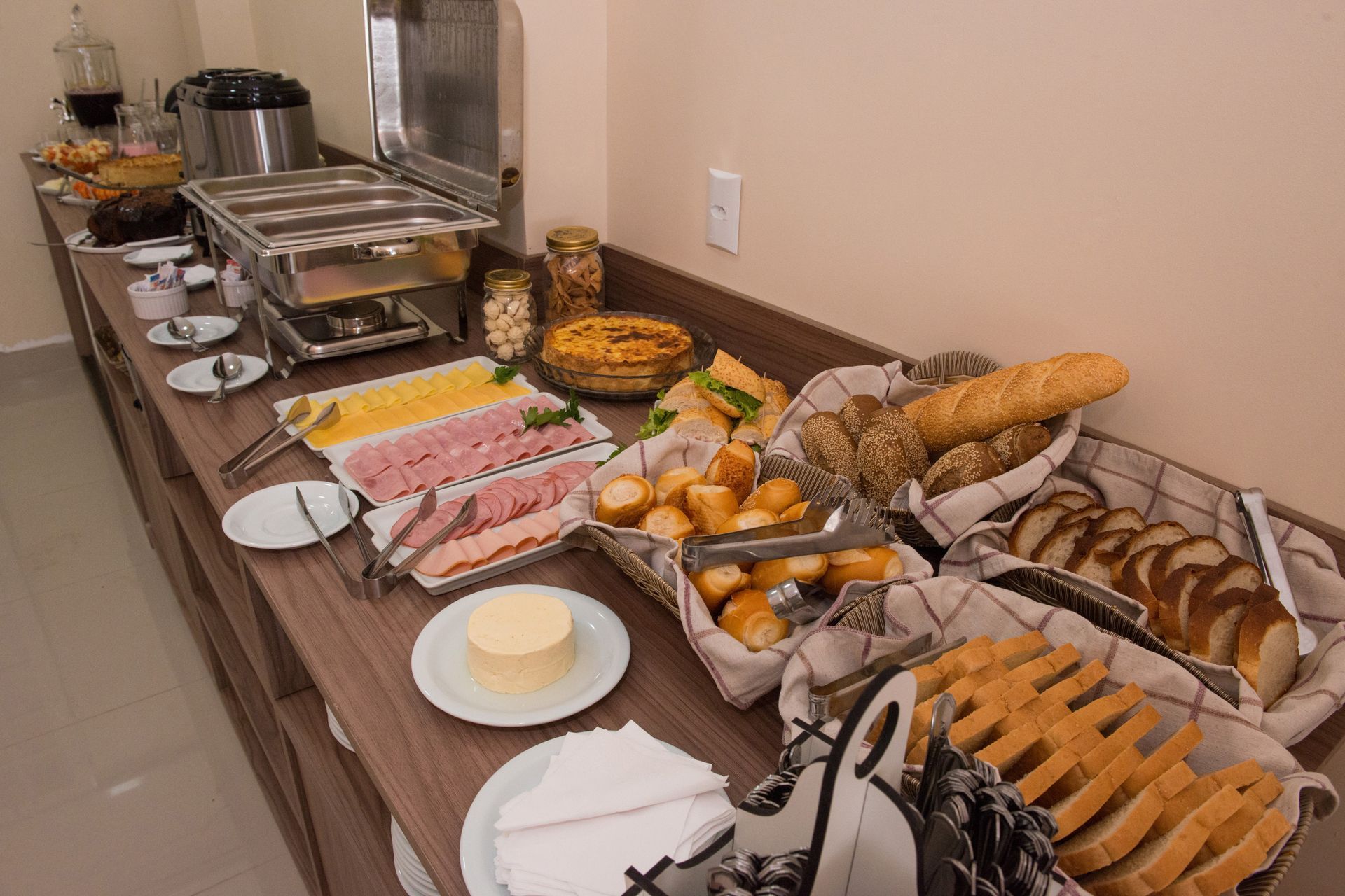Buffet de café da manhã com pães, frios, queijos, doces e comida quente aquecida.